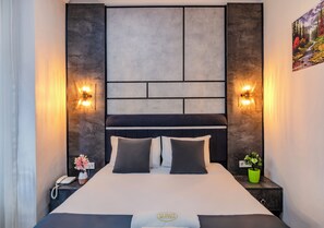 A modern hotel room with a large bed, a headboard with built-in lighting, a bedside table with a phone and a vase of flowers, and a small green plant on the nightstand.