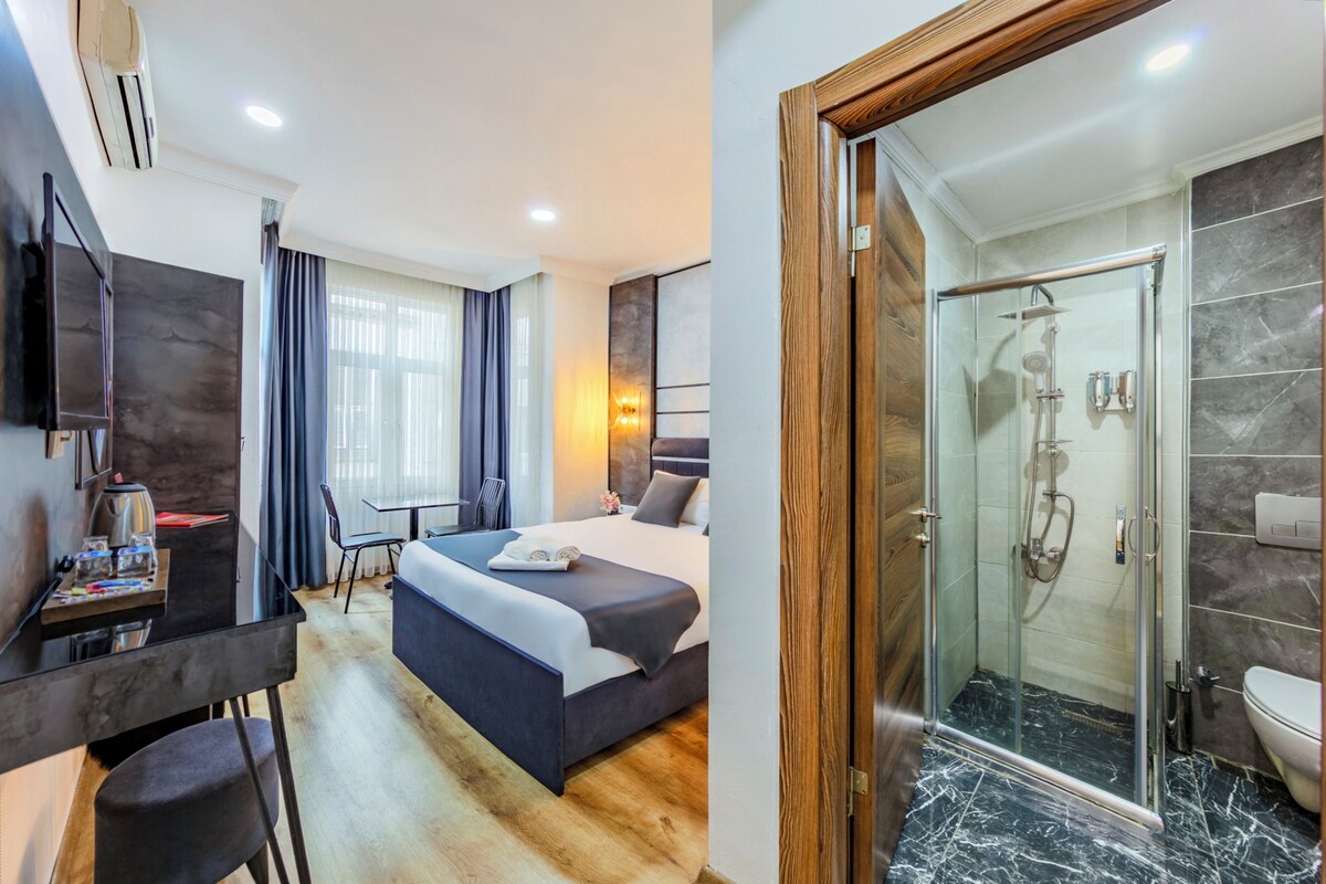 Una camera d'albergo con un letto, una scrivania, una sedia e un angolo doccia con porta in vetro.