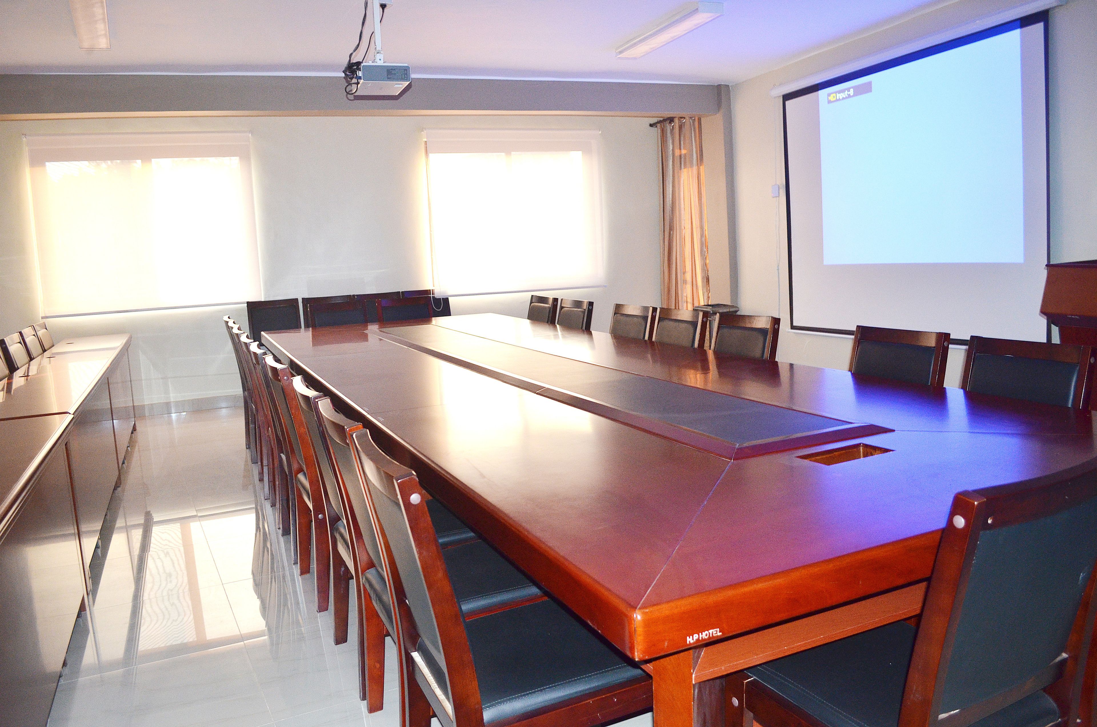 A conference room with a long table, chairs, a projector screen, and windows with blinds.
