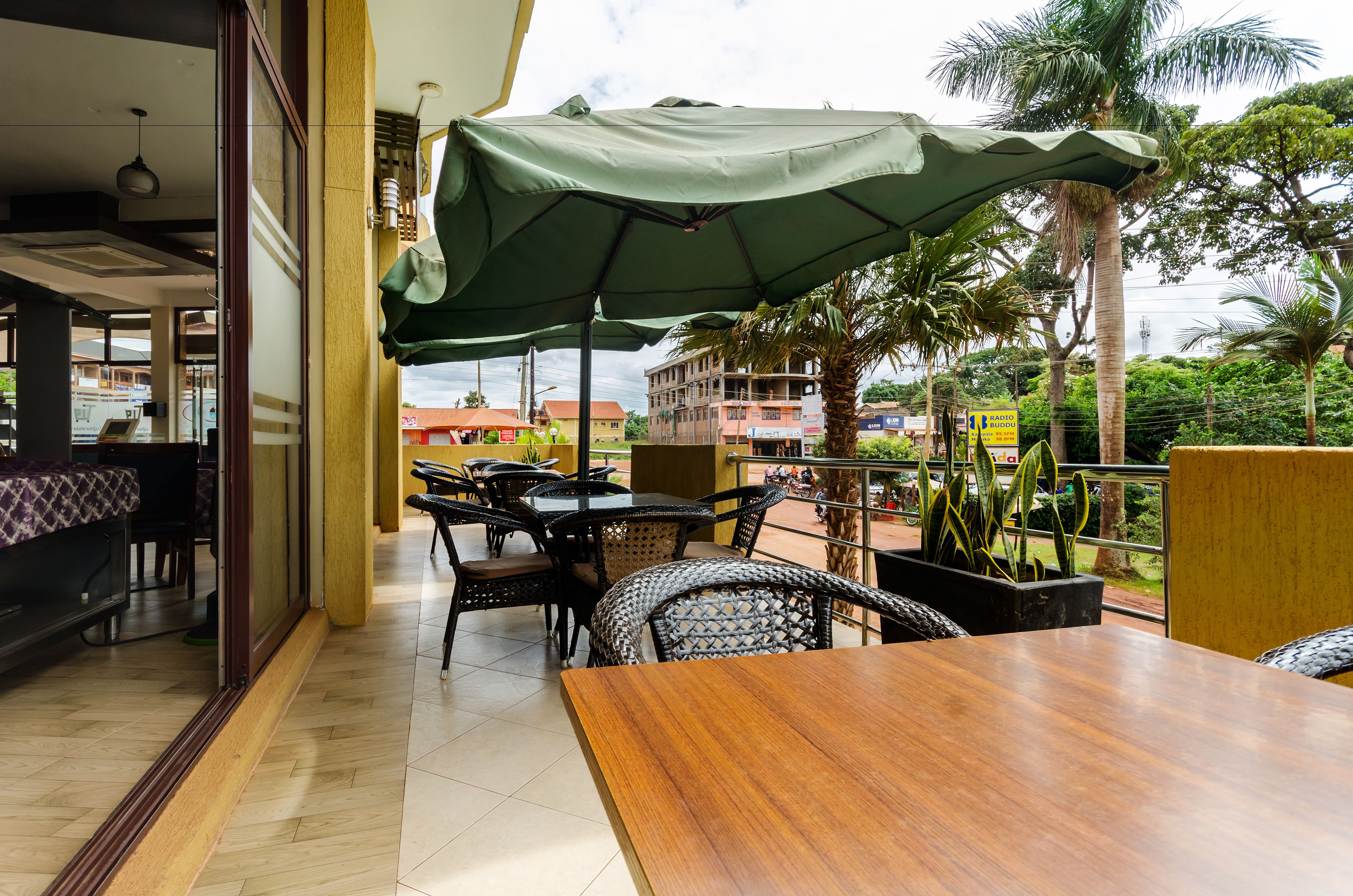 An outdoor dining area with wooden tables and chairs, a green umbrella, and a view of buildings and palm trees.