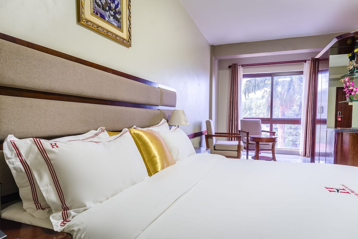 A hotel room with a large bed, a wooden headboard, a desk with a chair, and a window with curtains.