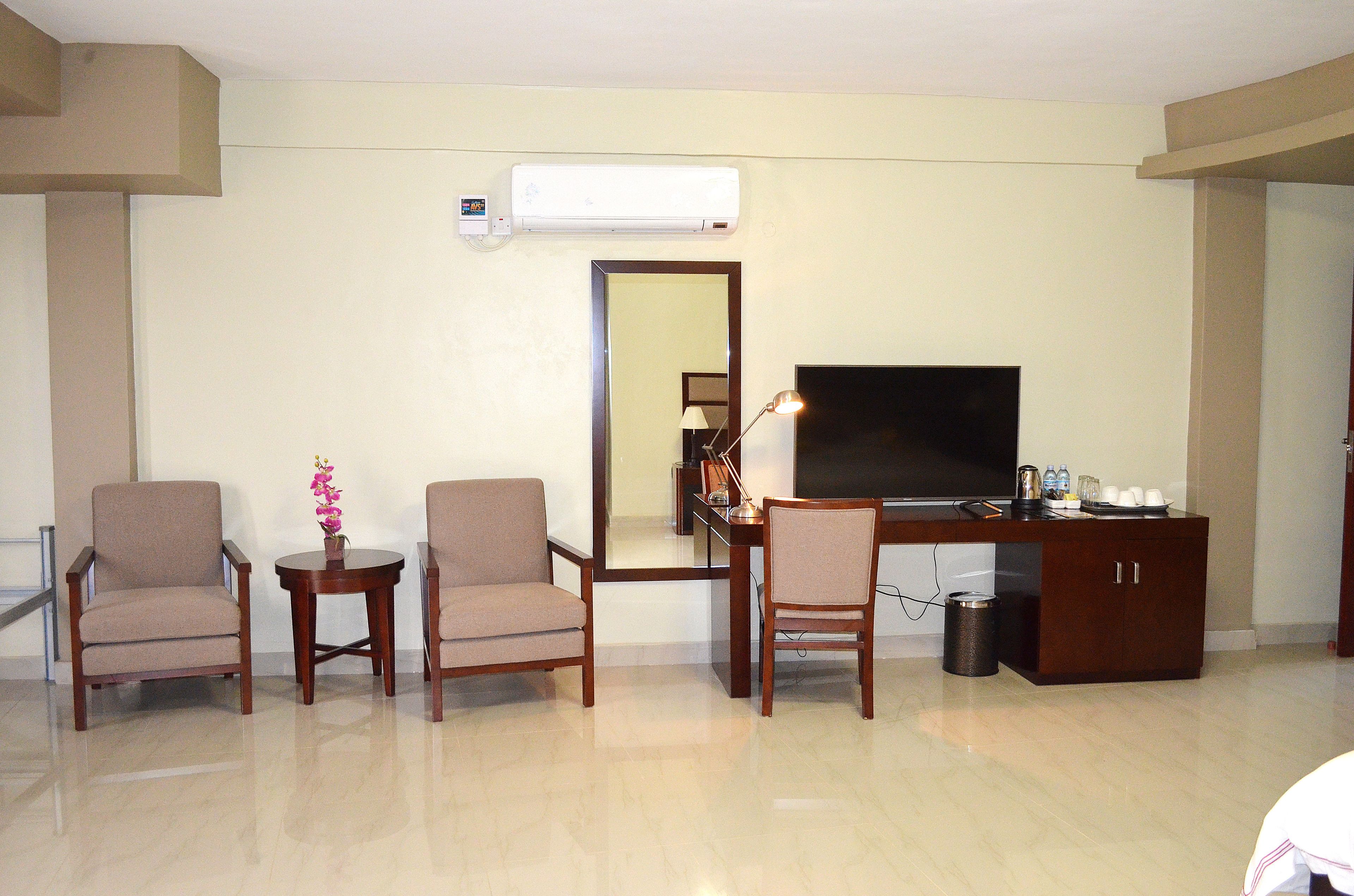 Habitación de hotel con televisión de pantalla plana, dos sillones, una mesita con un florero, un espejo y un armario de madera.