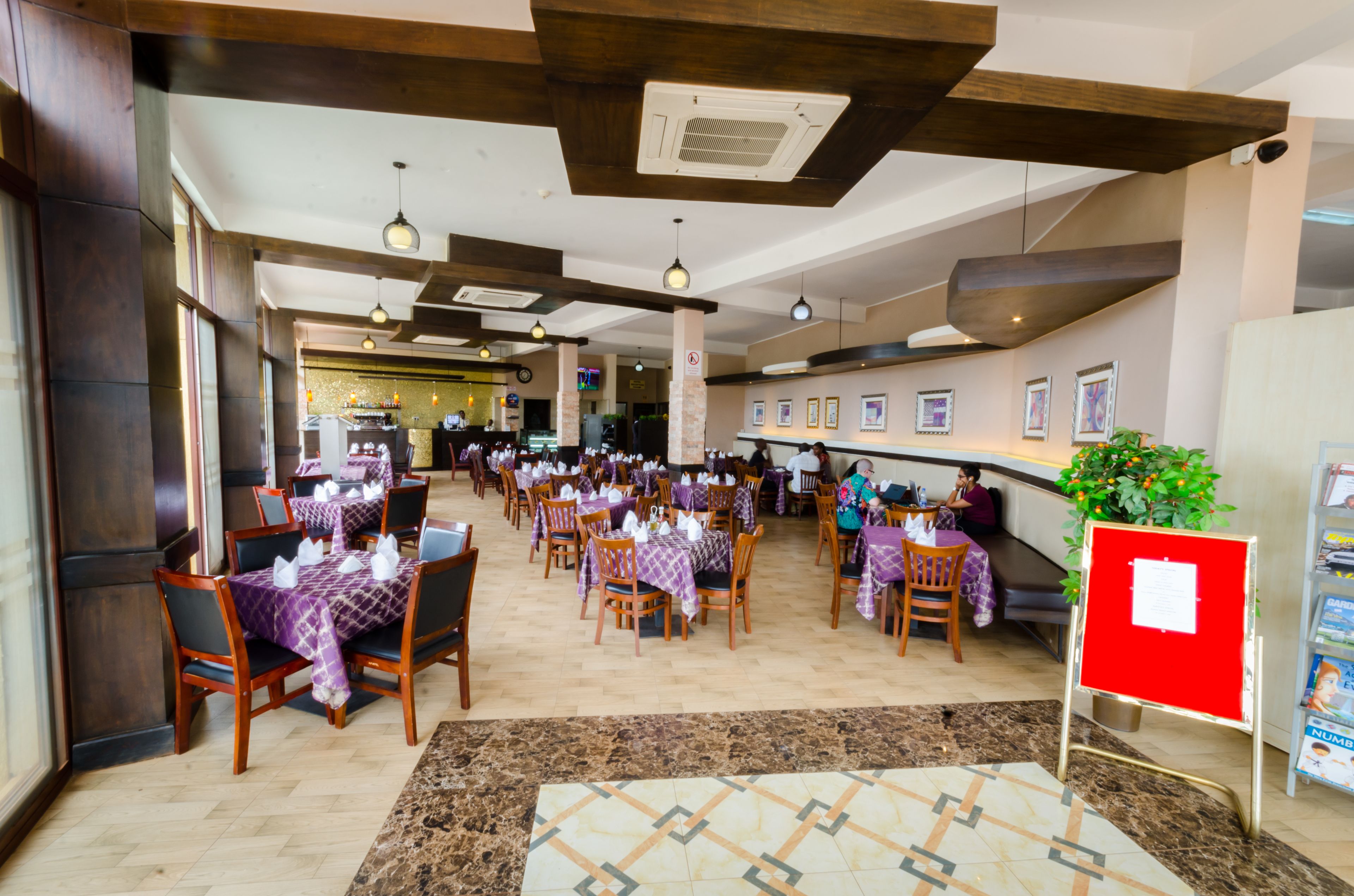 A spacious dining area with multiple tables and chairs, a central walkway, and a reception desk with framed pictures on the wall.