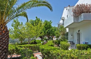 A white building with a balcony, surrounded by lush greenery and a palm tree.