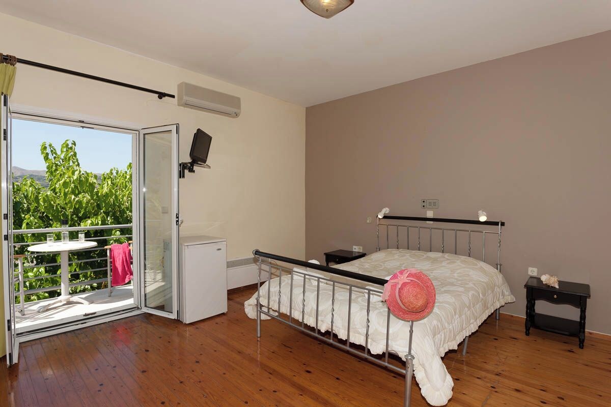 A bedroom with a bed, a small bedside table, a nightstand, a window with a view of greenery, and a sliding door to an outdoor area.