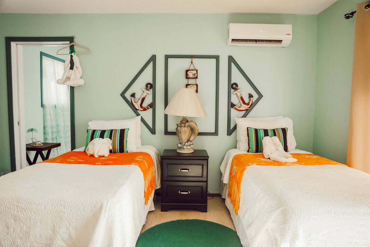 A hotel room with two beds, a nightstand, a lamp, and a wall-mounted air conditioner.
