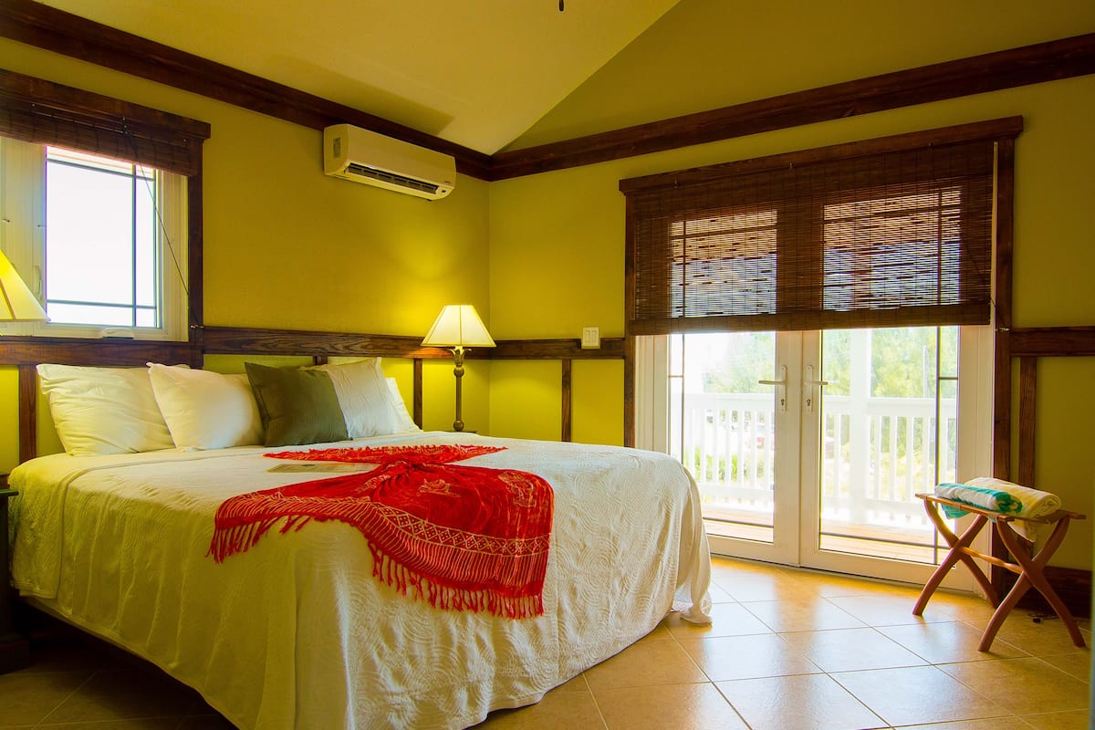 A hotel room with a bed, a window with blinds, an air conditioning unit, and a balcony with a view.
