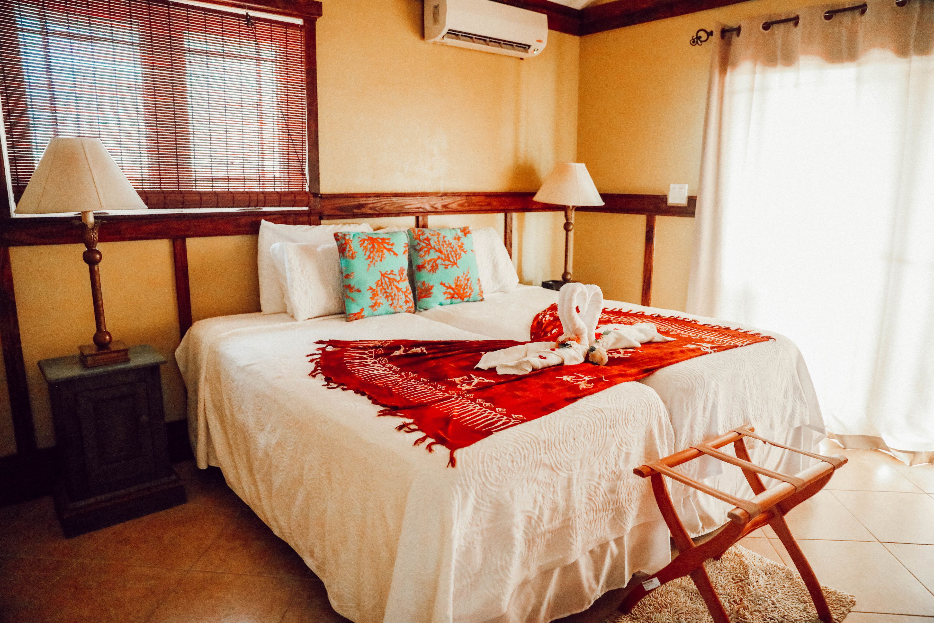 Una cama bien hecha con sábanas blancas, una manta roja y almohadones decorativos. Junto a la cama, una mesita de noche de madera con una lámpara.