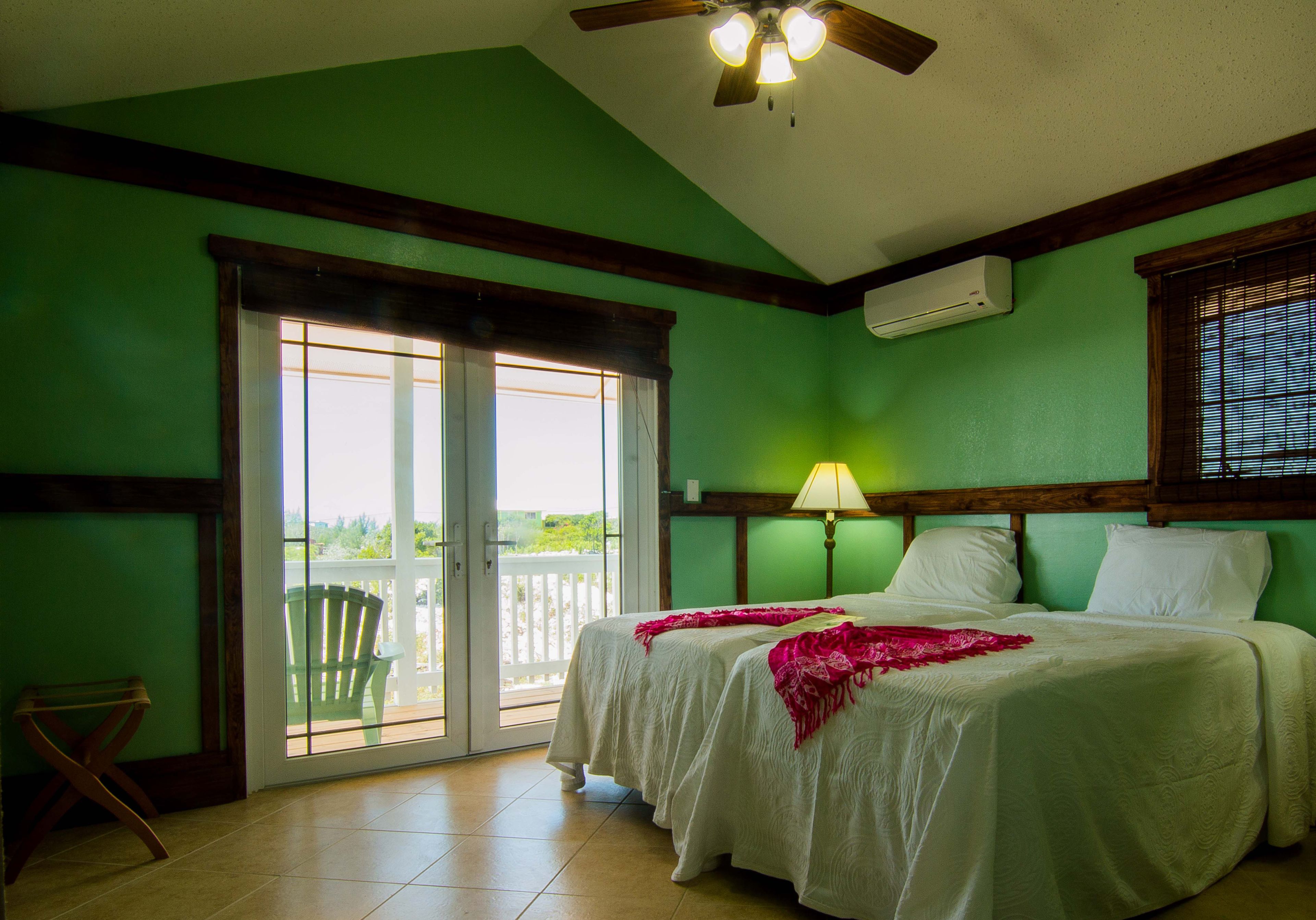 A room with two beds, a ceiling fan, a window with blinds, and a view of greenery outside.