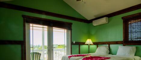 A room with two beds, a ceiling fan, a window with blinds, and a view of greenery outside.