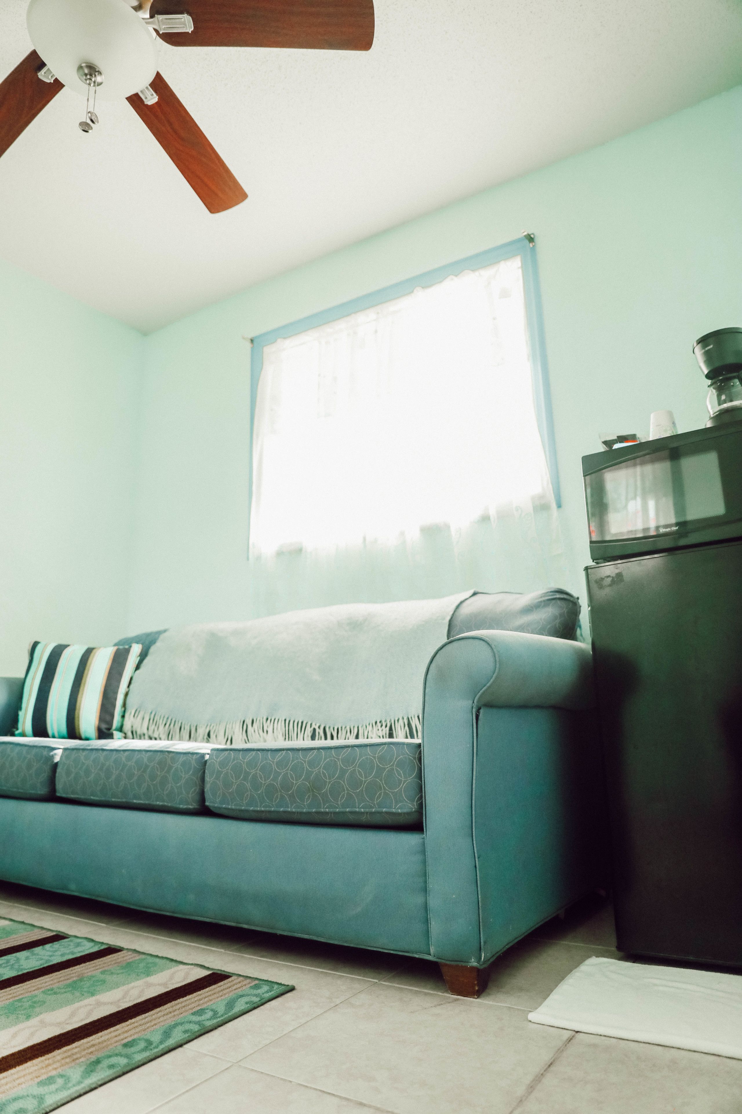 A room with a teal sofa, a ceiling fan, a window with curtains, and a refrigerator.