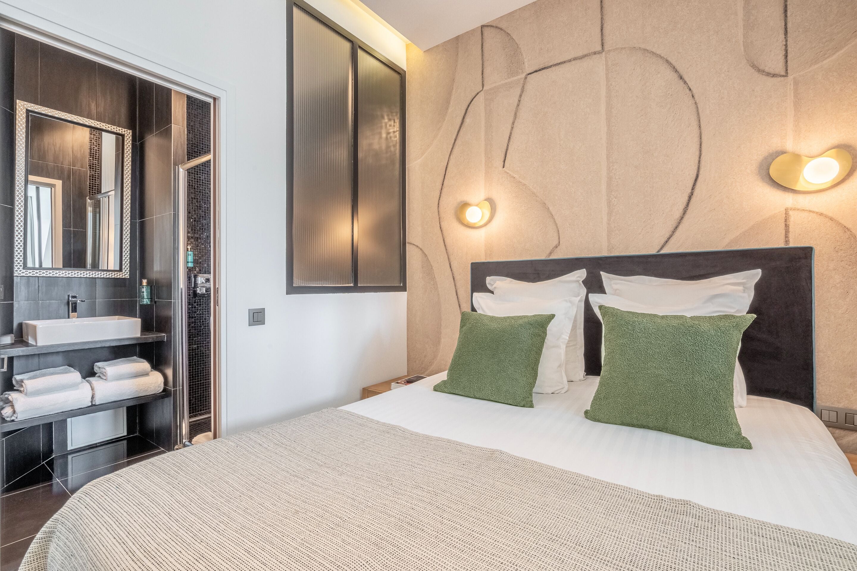 A modern bedroom with a large bed, two pillows, a nightstand, a mirror, and a bathroom with a sink and towel rack.