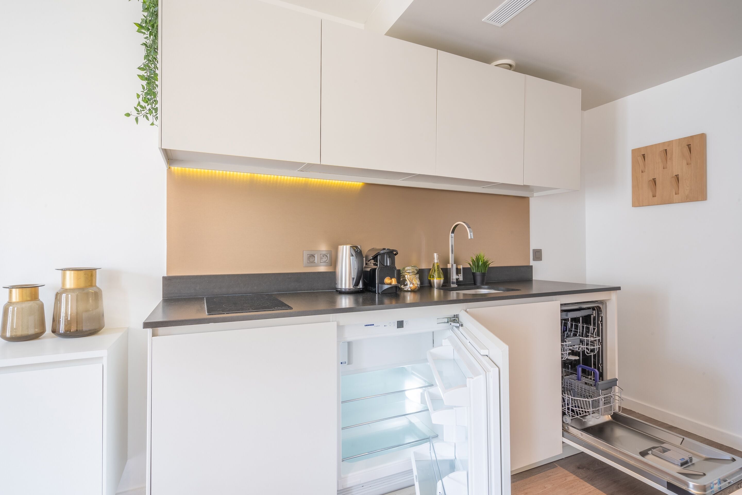 Une cuisine moderne avec des meubles blancs, un lave-vaisselle encastré et un plan de travail sur lequel se trouvent une bouilloire et une machine à café.