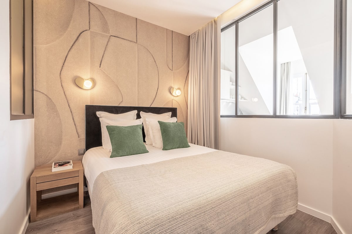 Moderni hotellihuone, jossa on suuri sänky, kaksi yöpöytävalaisinta, yöpöytä ja suuri ikkuna, josta avautuu näkymä kaupunkiin.