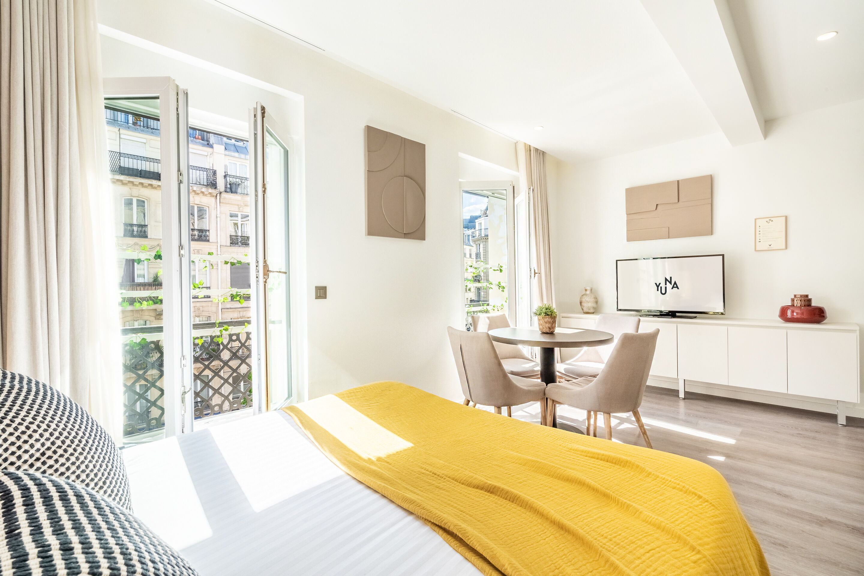 A hotel room with a bed, a dining area with chairs, a TV, and a balcony with plants.