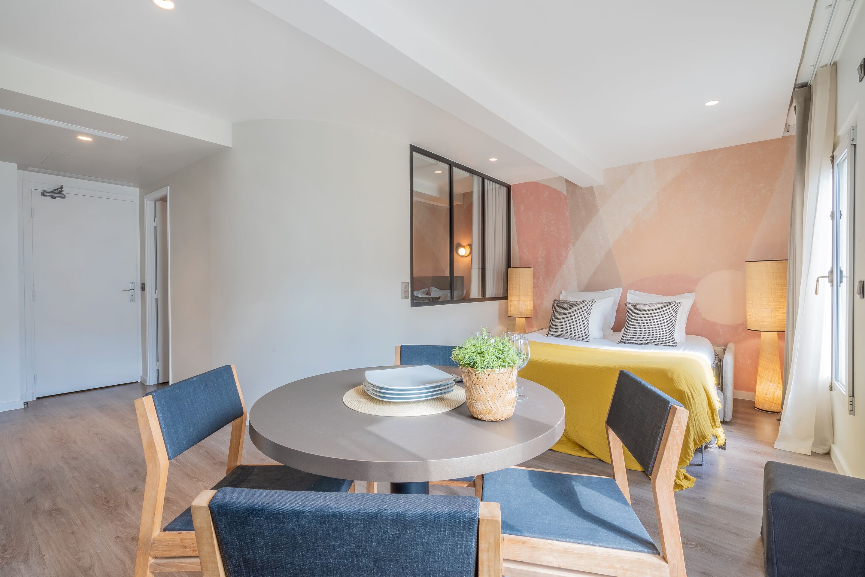 A modern hotel room with a round dining table, blue upholstered chairs, a bed with a yellow bedspread, and a large mirror on the wall.