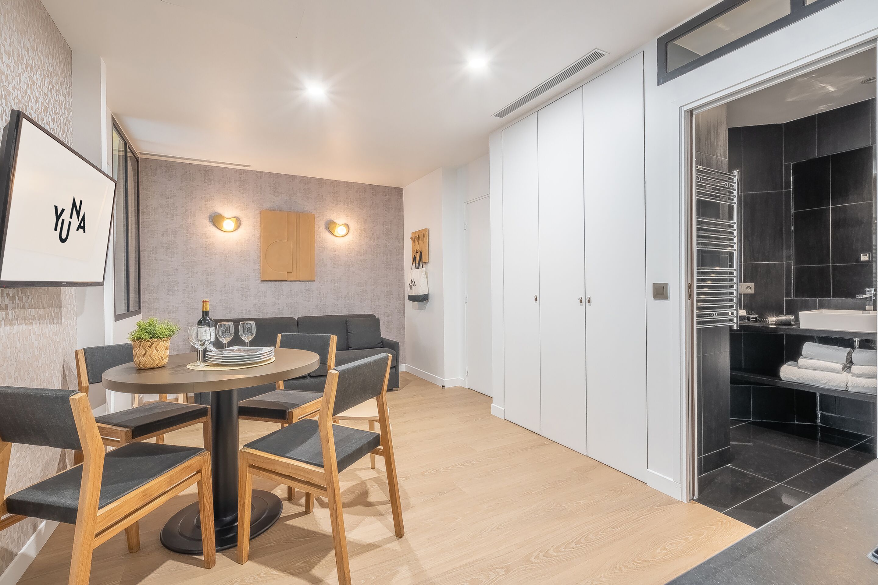 Un comedor moderno con una mesa redonda, sillas y un sofá. Cuenta con una zona de cocina con salpicadero de azulejos negros y armarios blancos.