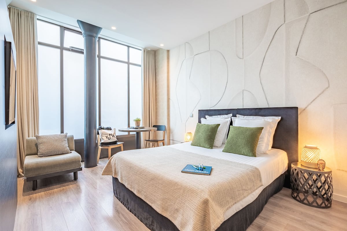 Moderni hotellihuone, jossa on suuri sänky, pieni työpöytä, tuoli ja suuret ikkunat, joista avautuu näkymä kaupunkiin.