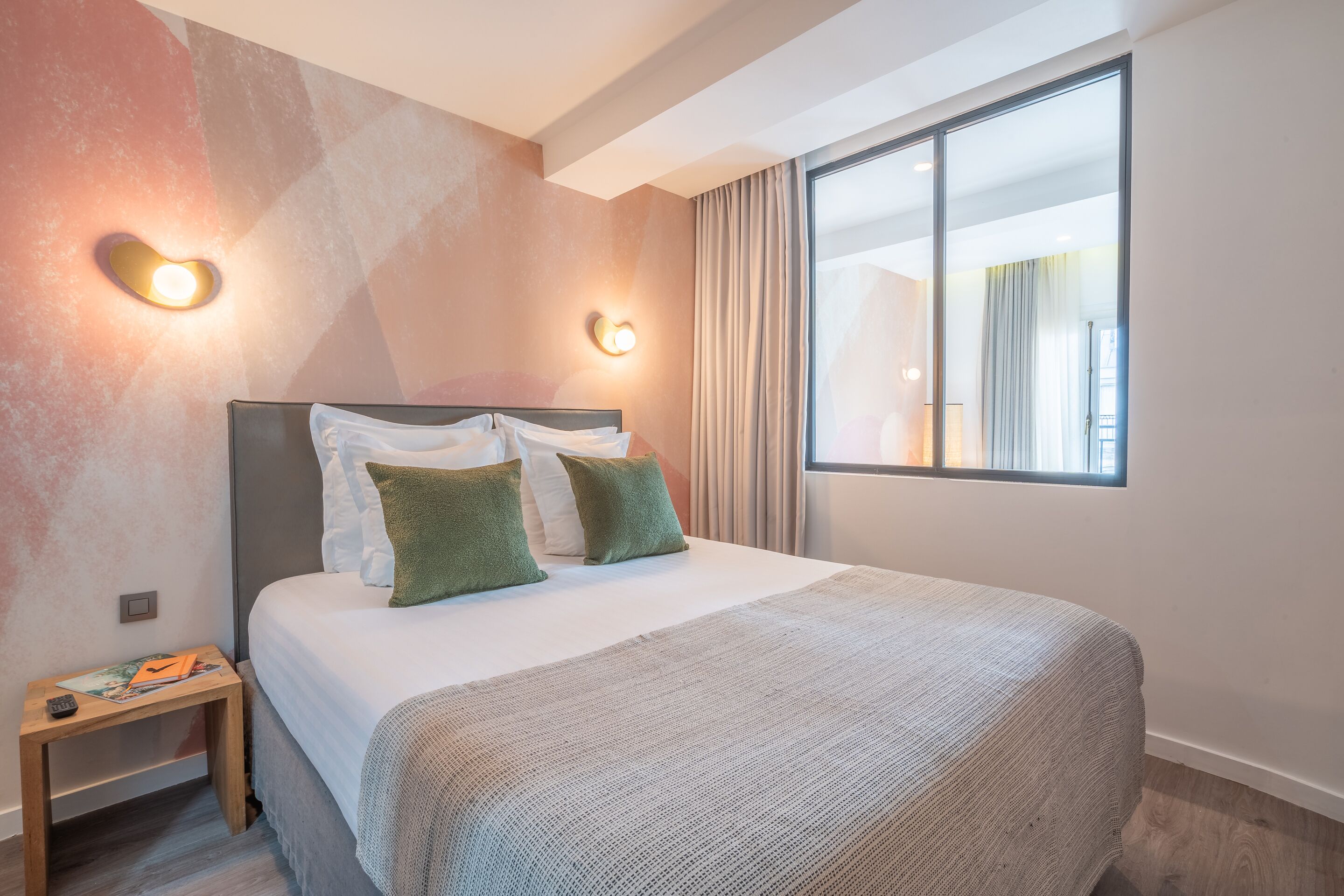 A hotel room with a large bed, two wall sconces, a nightstand, and a window with curtains.