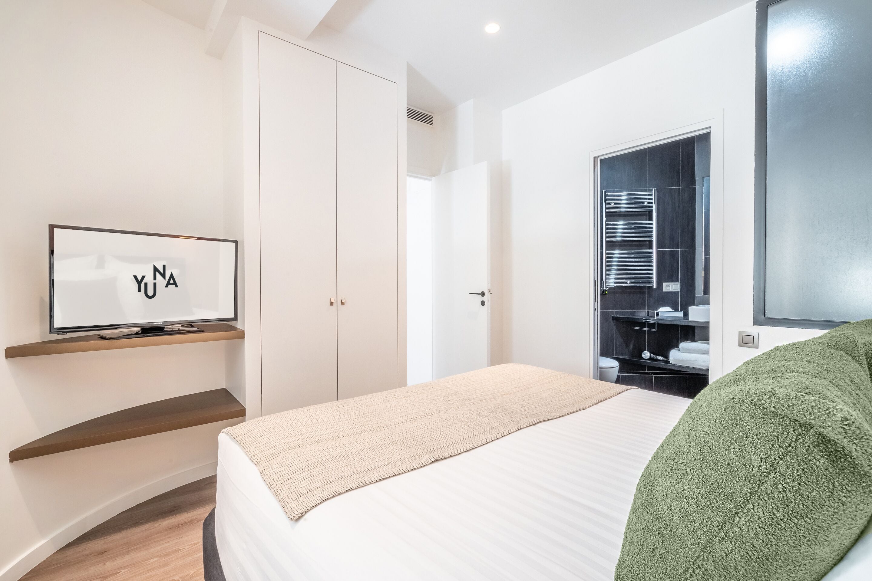 A modern hotel room with a bed, a TV mounted on a wall, a wardrobe, and a bathroom with a mirror and towel rack.