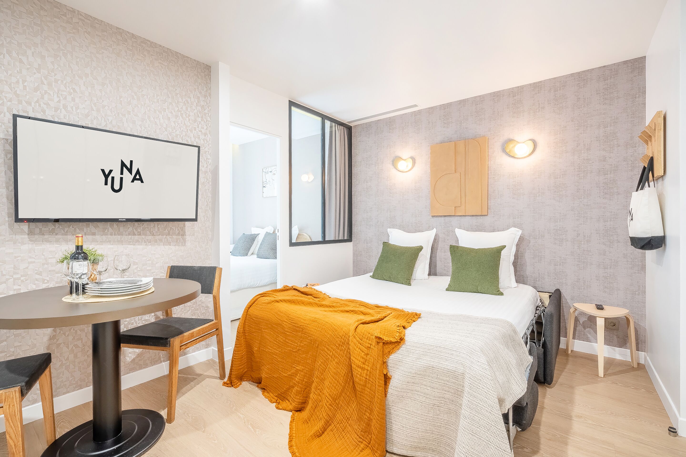 A modern hotel room with a bed, a round dining table, and a TV mounted on the wall.