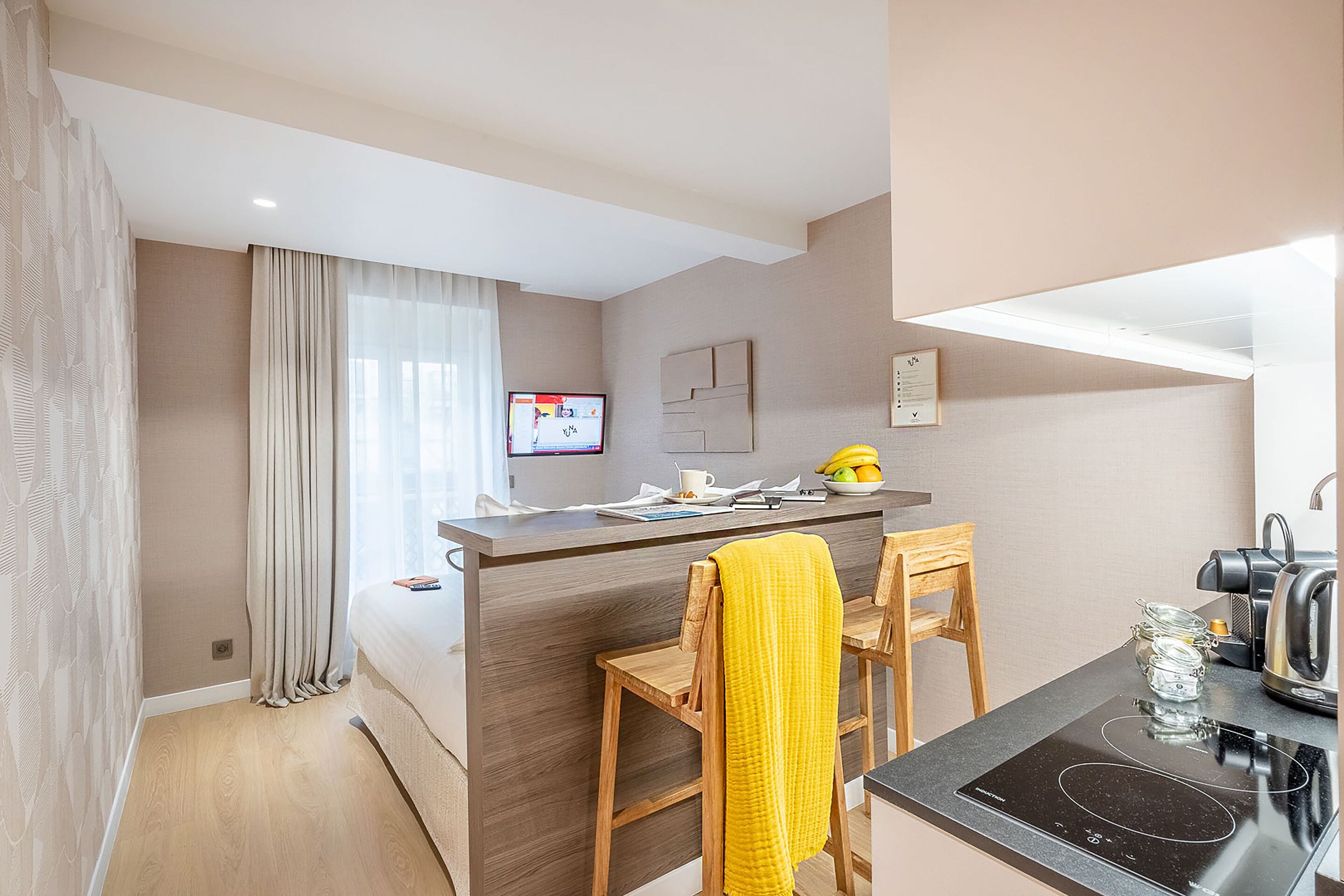 A modern kitchen with a central island, wooden chairs, a yellow towel, a kettle, and a TV on the wall.
