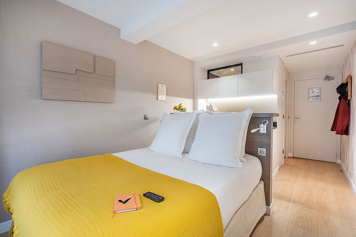 Hotellihuone, jossa on sänky, yöpöytä, peili, ovi ja seinään kiinnitetty valaisin.