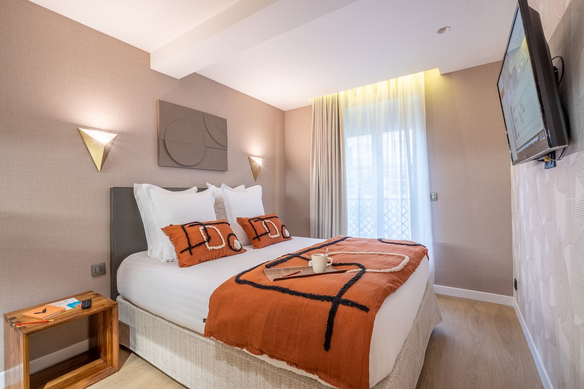 Moderni hotellihuone, jossa on suuri sänky, yöpöytä, seinälle asennettu televisio ja ikkuna, jossa on verhot.