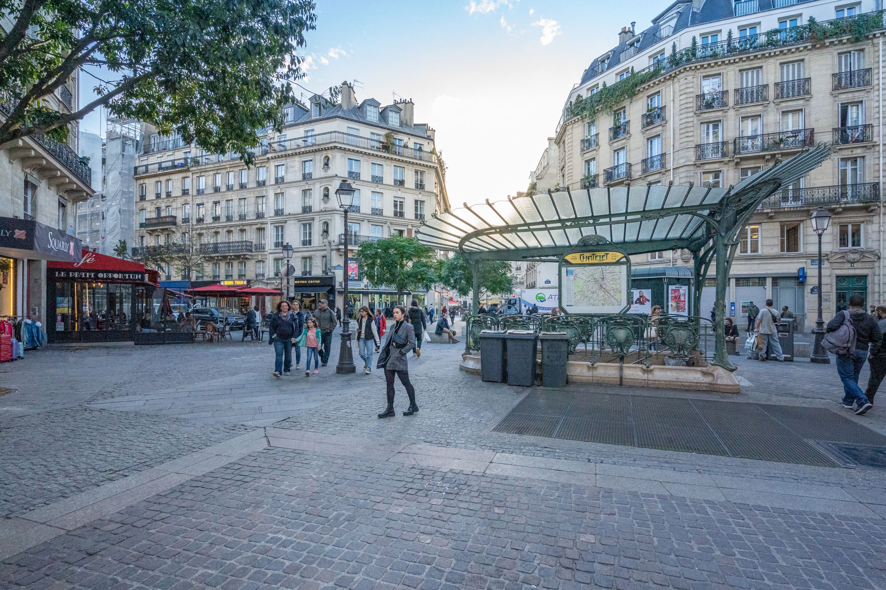 A public square with cobblestone pavement, a covered seating area, and surrounding buildings with commercial establishments.