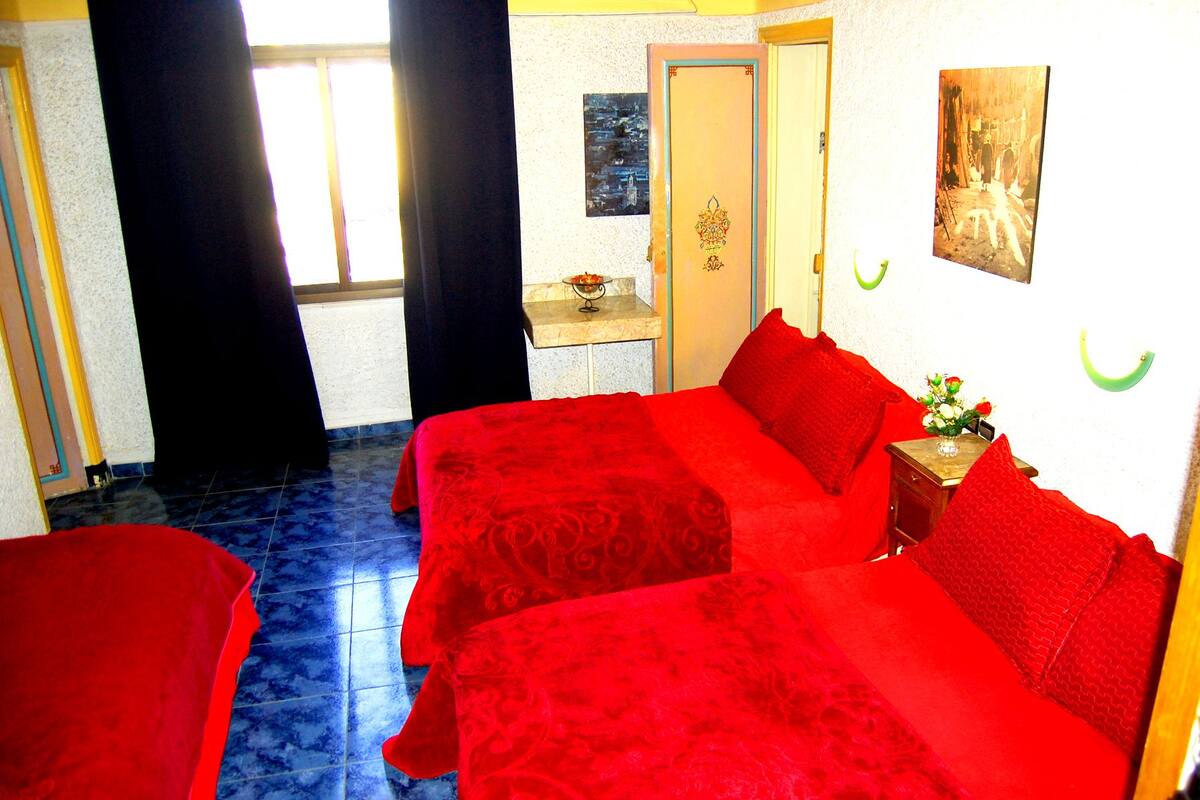 A hotel room with two red beds, a small table with a vase, and a painting on the wall.