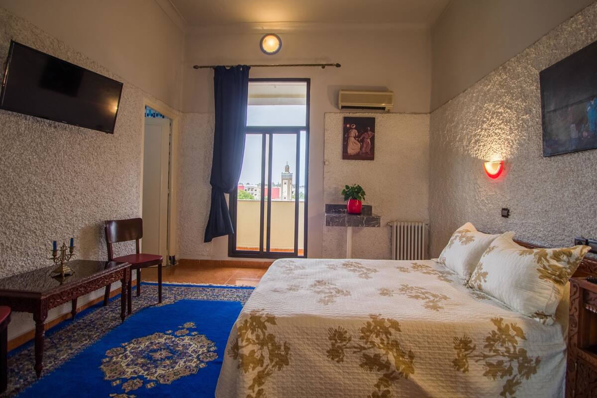 A bedroom with a bed, a desk, a chair, a blue carpet, and a view through a window.