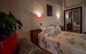 Un dormitorio con cama, una mesita de noche, un jarrón rojo con planta e iluminación en la pared.
