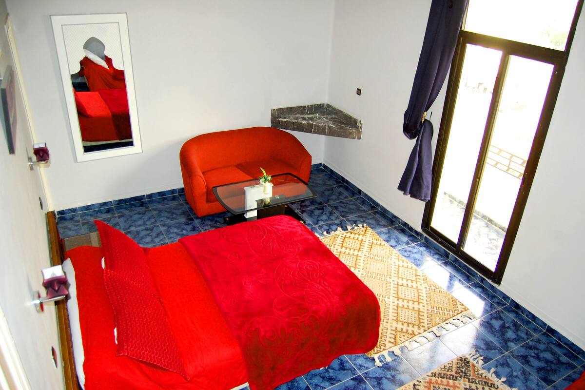 A room with a red sofa, a glass coffee table, a patterned rug, a framed artwork on the wall, and a window with curtains.