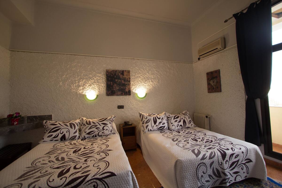 A room with two beds, a nightstand, a wall-mounted heater, and a window with curtains.