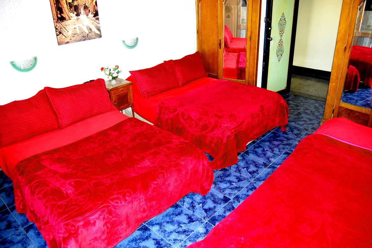 A room with two red beds, a small table with a vase of flowers, and a mirror on the wall.