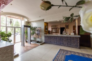Un vestíbulo de hotel con una recepción, un muro de azulejos azules y blancos con diseño, un ventanal con vistas a árboles y una alfombra azul.