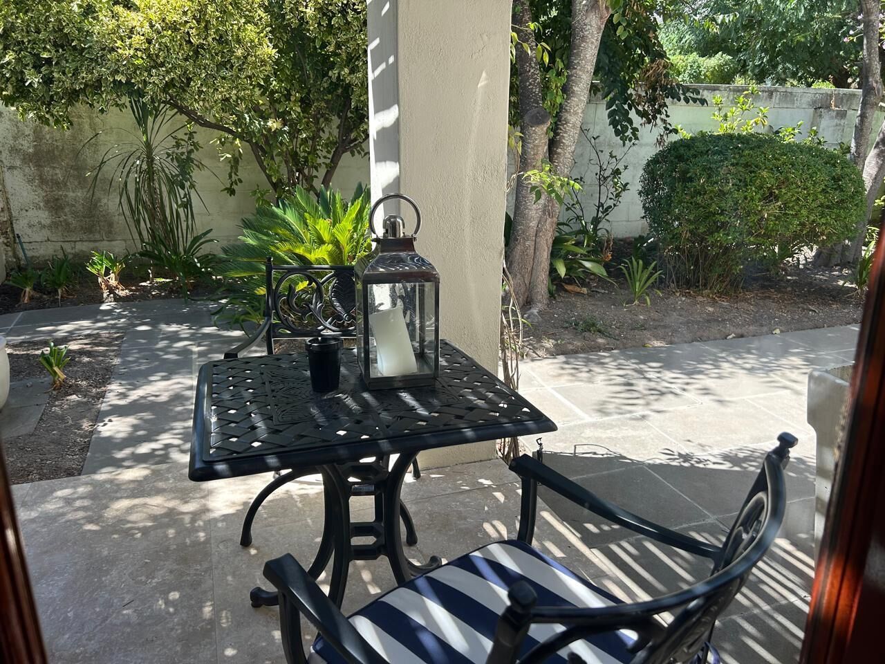 An outdoor patio with a metal table and chairs, surrounded by greenery and a concrete wall.