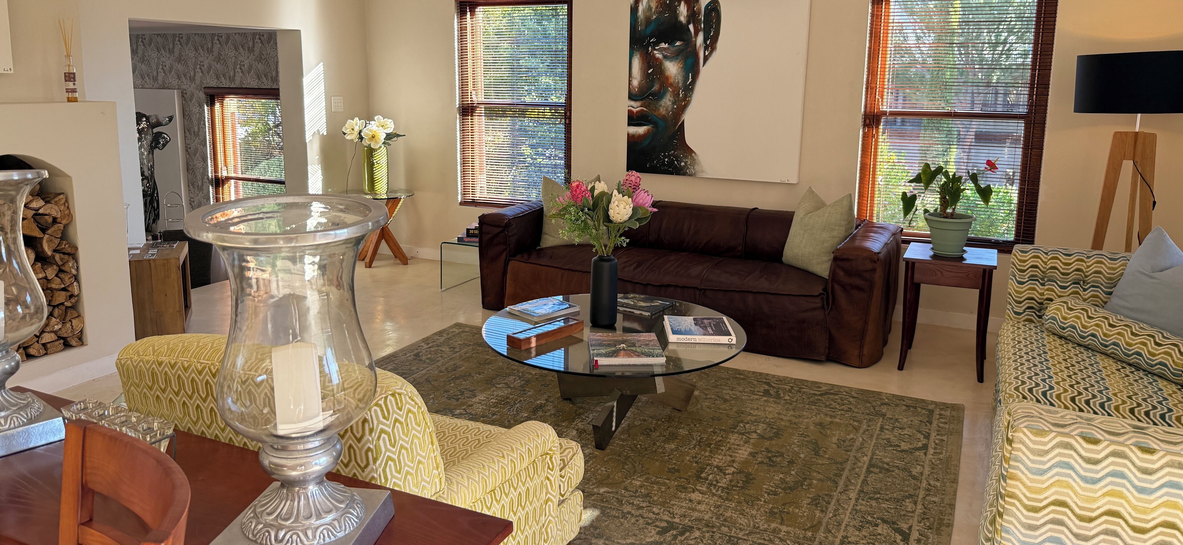A living room with a brown sofa, a glass coffee table, and a patterned rug. There are two windows with blinds, a painting on the wall, and a potted plant on a side table.