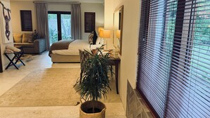 A spacious bedroom with a large bed, a sitting area with a sofa, and a dining table with chairs. There is a window with blinds and a view of greenery outside. - Residence Vive La Vie (Franschhoek)