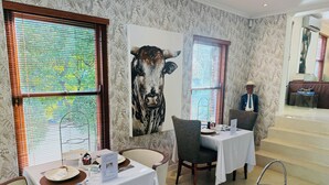 A restaurant with two tables, each with a white tablecloth and a menu stand. There is a large painting of a cow on the wall, and a person in a suit is visible in the background. - Residence Vive La Vie (Franschhoek)