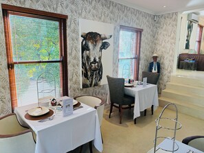 Un restaurant avec deux tables, chacune recouverte d’une nappe blanche et agrémentée d’un présentoir à menus. Un grand tableau représentant une vache est accroché au mur, et on aperçoit une personne en costume à l’arrière-plan.