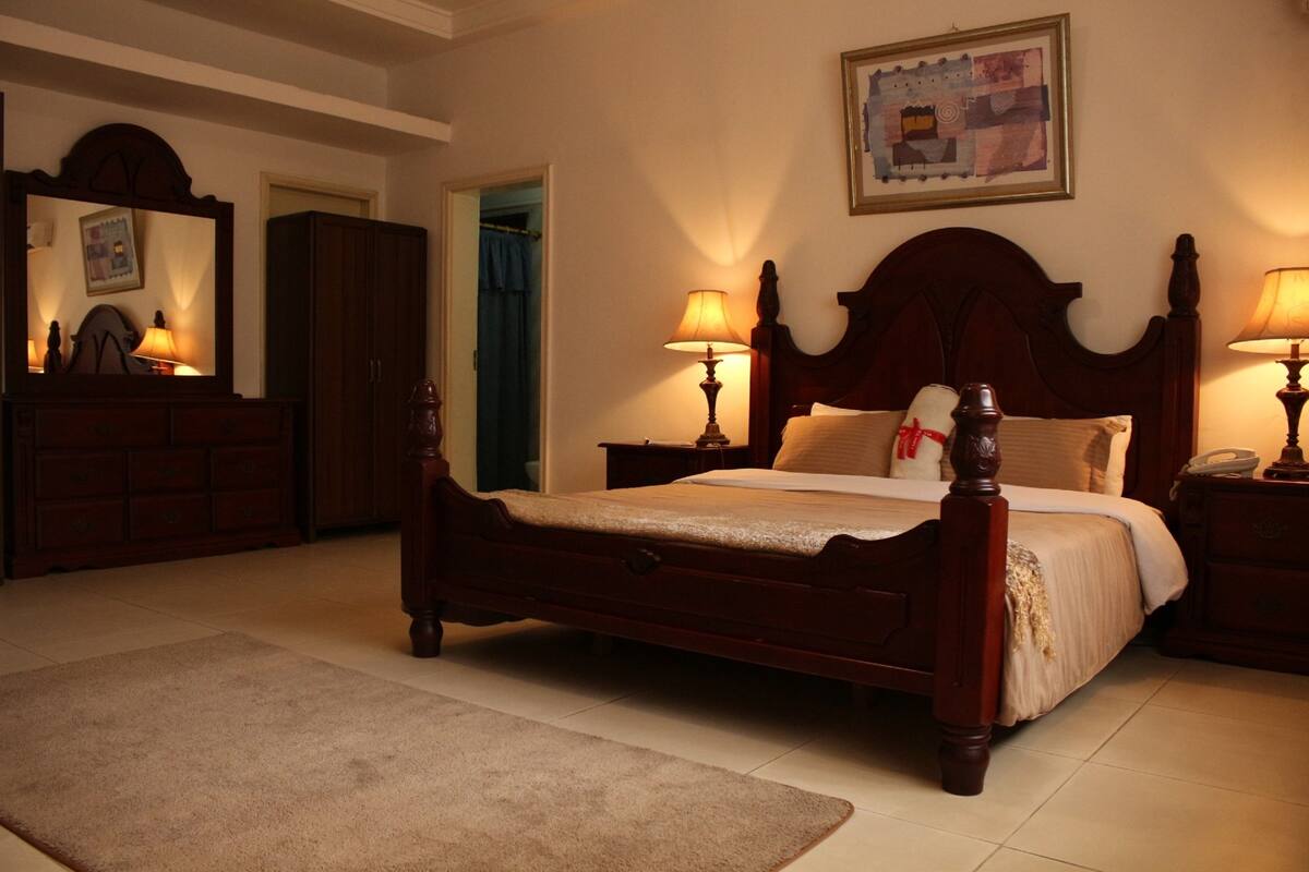 A traditional bedroom with a four-poster bed, a wooden dresser, and a nightstand with a lamp.