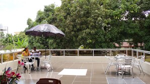 Un patio al aire libre con sillas y mesas de metal, un paraguas grande y vistas a árboles y edificios.