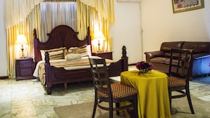 Un dormitorio con una cama, dos mesitas de noche con lámparas, una silla, una pequeña mesa redonda con mantel amarillo y un sofá de cuero marrón.
