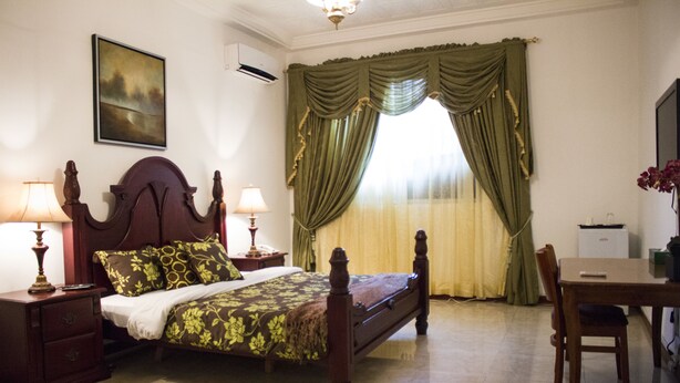Un dormitorio con una cama de madera, dos mesitas de noche con lámparas, una pequeña mesa con una silla y una ventana con cortinas verdes.