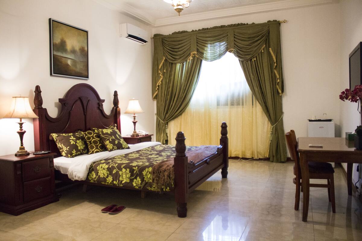 A bedroom with a wooden bed, two bedside tables with lamps, a small table with a chair, and a window with green curtains.