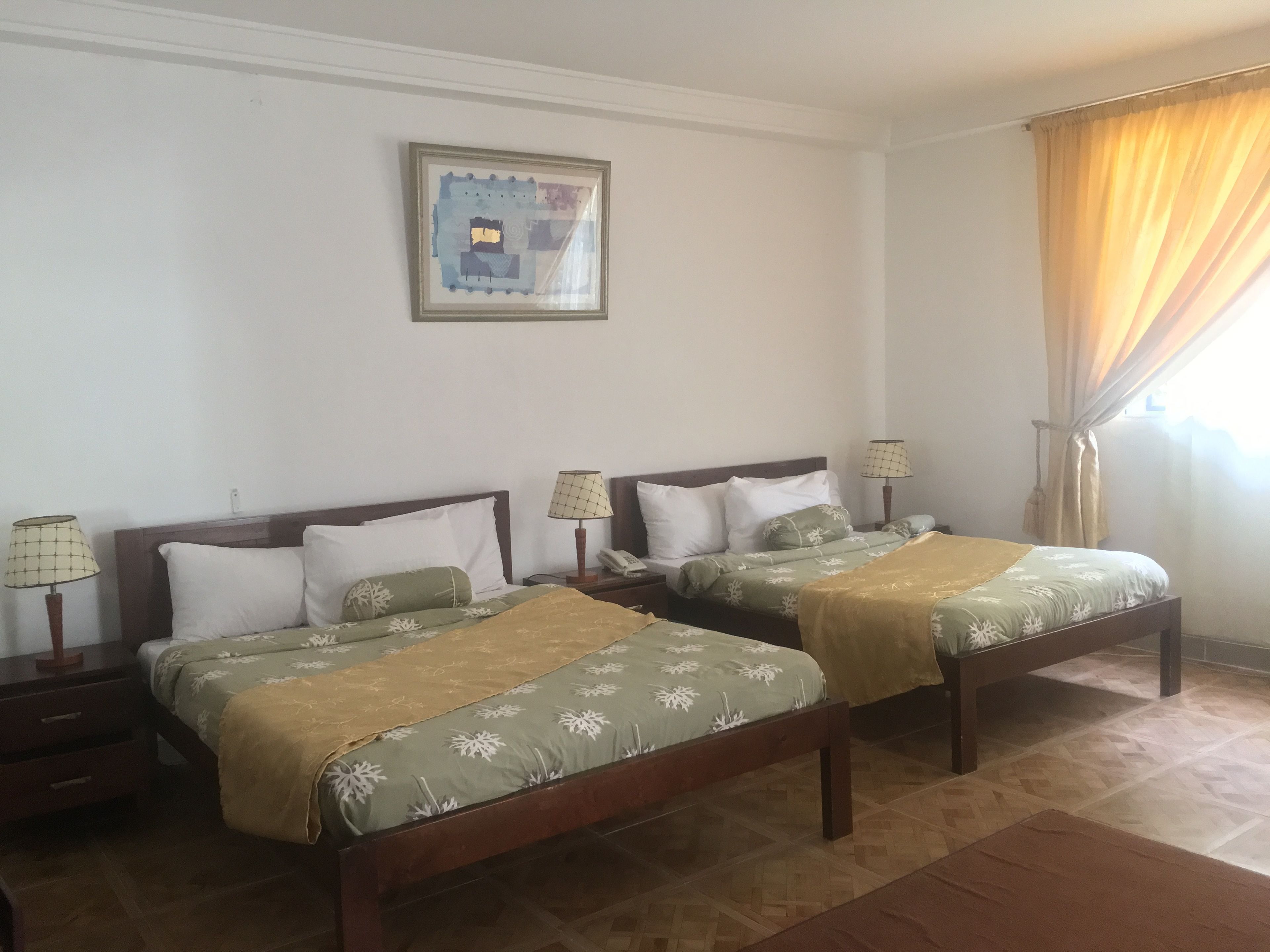 Ein Hotelzimmer mit zwei Betten, beide mit Holzrahmen und grüner Bettwäsche. Es gibt Nachttische mit Lampen, ein gerahmtes Bild an der Wand und ein Fenster mit Vorhängen.