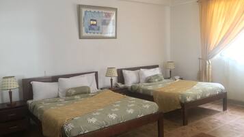 Ein Hotelzimmer mit zwei Betten, beide mit Holzrahmen und grüner Bettwäsche. Es gibt Nachttische mit Lampen, ein gerahmtes Bild an der Wand und ein Fenster mit Vorhängen.