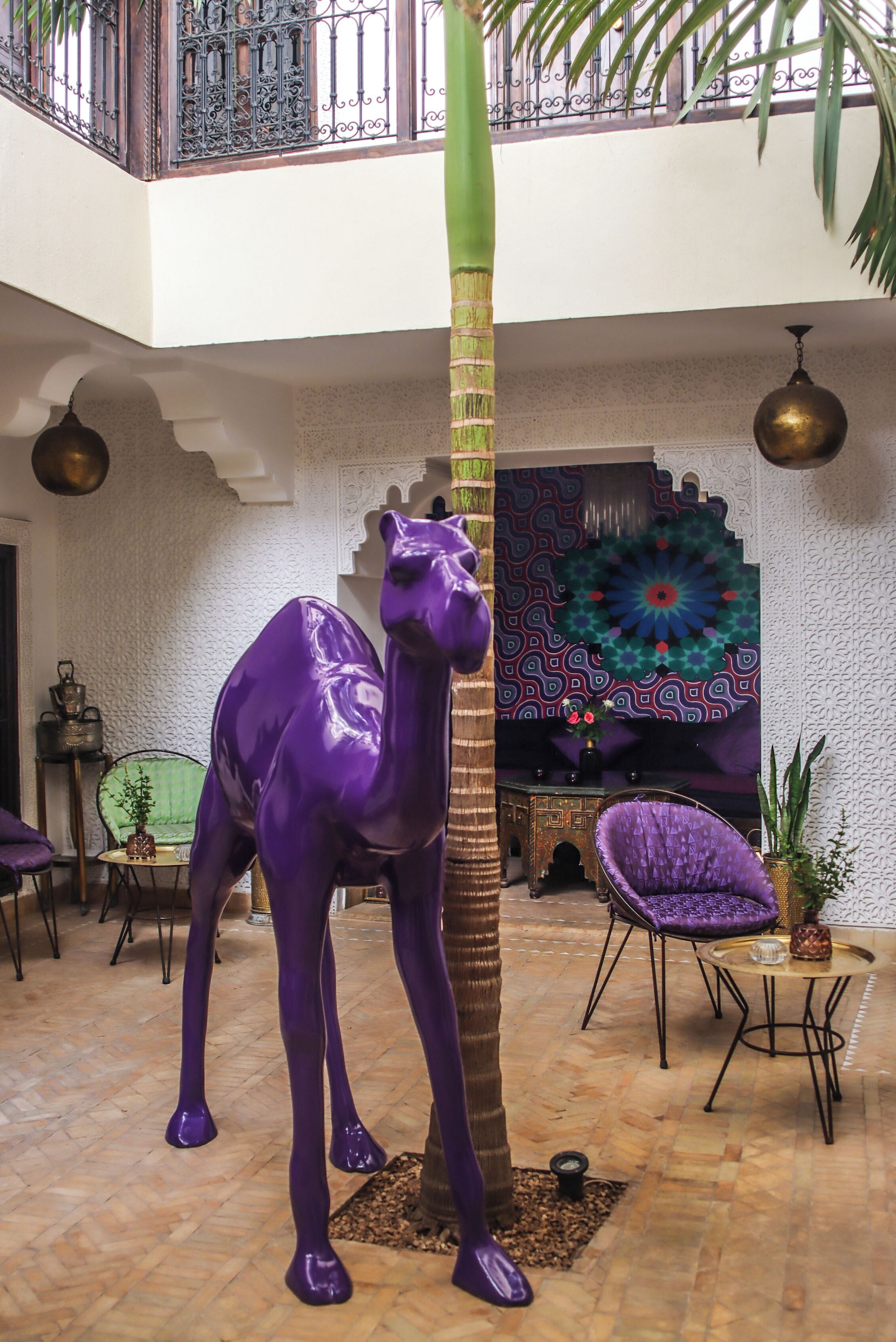A camel statue, a palm tree, and a patterned wall in a lobby area with a high ceiling and a balcony.