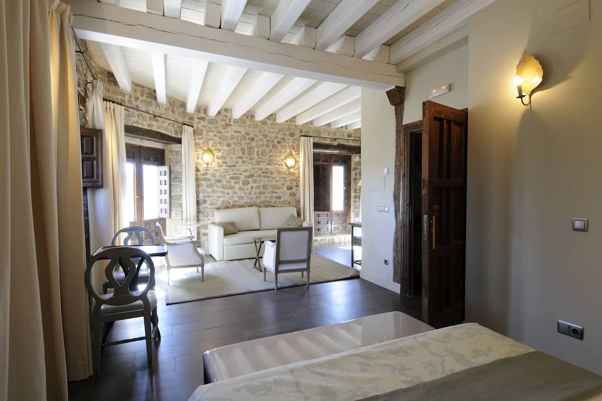 Una habitación con pared de piedra, vigas de madera, un sofá blanco, una mesa de comedor y una cama.